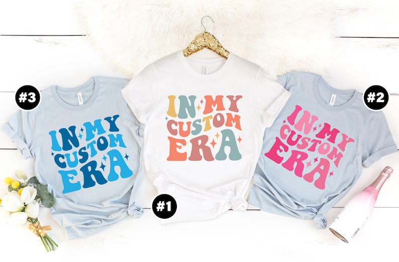 May include: Three t-shirts with the text "In My Custom Era" in a retro font with stars and different color schemes. The shirts are light blue, white, and pink.