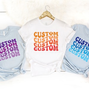 May include: Three t-shirts with the word "CUSTOM" repeated in a gradient pattern. The shirts are light blue, white, and light blue. The gradient pattern on the shirts is purple to pink, orange to red, and blue to light blue.
