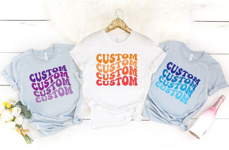 May include: Three t-shirts with the word "CUSTOM" repeated in a gradient pattern. The shirts are light blue, white, and light blue. The gradient pattern on the shirts is purple to pink, orange to red, and blue to light blue.