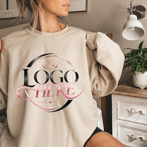 May include: Beige sweatshirt with the text "LOGO HERE" in a black and pink design. The logo features a crown and a circular design. The sweatshirt is a neutral color and has long sleeves.