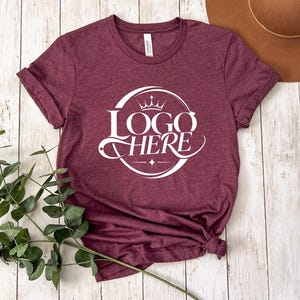 May include: Burgundy t-shirt with rolled sleeves. The shirt features a white circular logo with a crown and the words "LOGO HERE" in a stylized font. The shirt is knotted at the bottom. A brown hat is visible in the background.