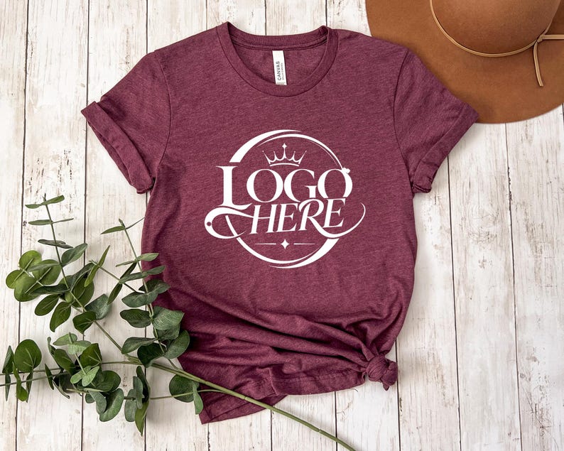 May include: Burgundy t-shirt with rolled sleeves. The shirt features a white circular logo with a crown and the words "LOGO HERE" in a stylized font. The shirt is knotted at the bottom. A brown hat is visible in the background.