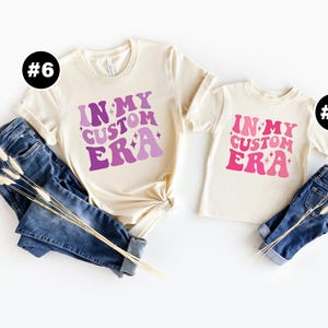 May include: Two cream colored t-shirts with the text "In My Custom Era" in pink and purple lettering. The shirts are laid out on a white background with blue jeans and dried flowers.