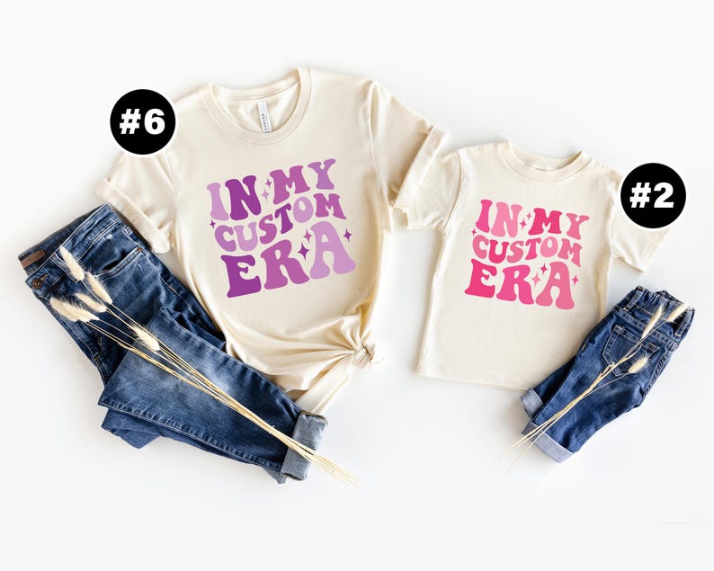 May include: Two cream colored t-shirts with the text "In My Custom Era" in pink and purple lettering. The shirts are laid out on a white background with blue jeans and dried flowers.