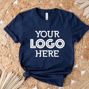 May include: Navy blue t-shirt with the text "YOUR LOGO HERE" in white. The shirt is knotted at the bottom and is displayed on a woven, circular rug. The shirt has rolled-up sleeves.