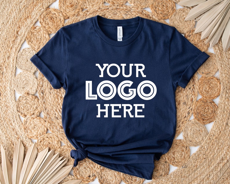 May include: Navy blue t-shirt with the text "YOUR LOGO HERE" in white. The shirt is knotted at the bottom and is displayed on a woven, circular rug. The shirt has rolled-up sleeves.