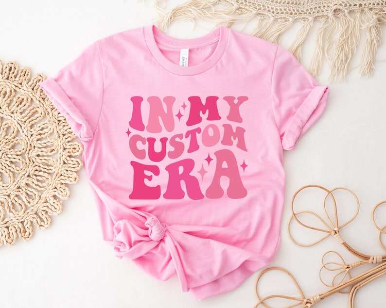 May include: Pink t-shirt with the text "IN MY CUSTOM ERA" in a retro font with pink and white glitter accents.