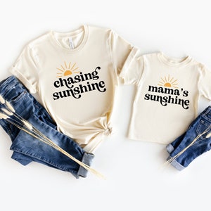 May include: Two beige t-shirts with a sun graphic and text. The larger shirt says "chasing sunshine" and the smaller shirt says "mama's sunshine".