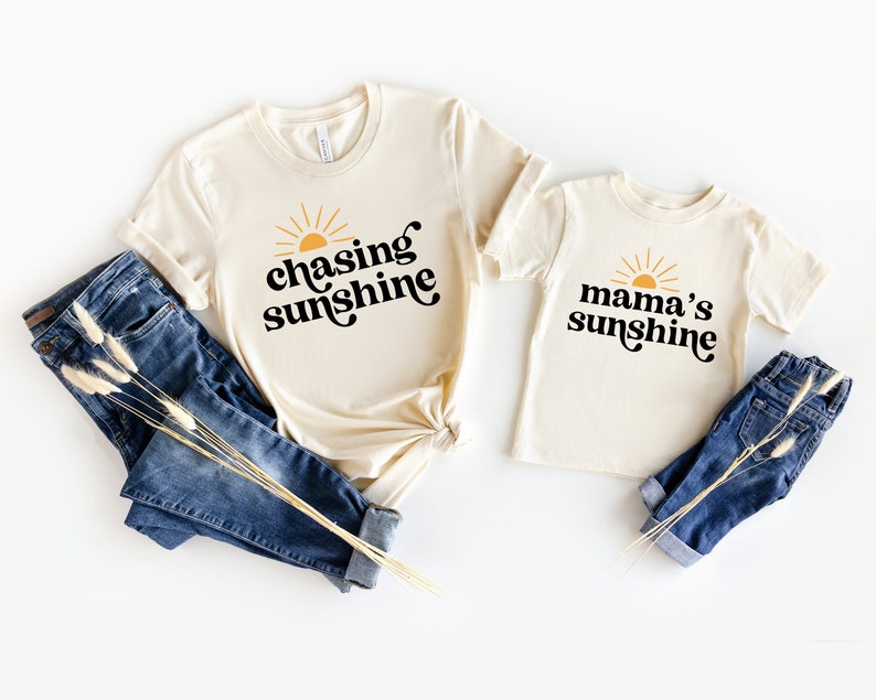 May include: Two beige t-shirts with a sun graphic and text. The larger shirt says "chasing sunshine" and the smaller shirt says "mama's sunshine".