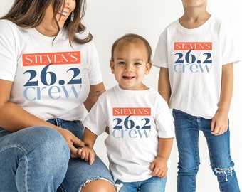 May include: Three people wearing white t-shirts with the text "STEVEN'S 26.2 CREW" in red, blue, and light blue. The people are wearing blue jeans and white shoes. The image is taken in front of a white background.