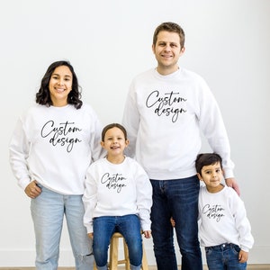 May include: A family of four wearing white sweatshirts with the text "Custom Design" in black script. The family is standing in front of a white wall.