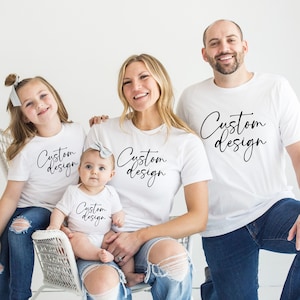 May include: A family of four wearing white t-shirts with the text "Custom Design" in black script. The family is sitting on a white chair and the floor. The adults are wearing blue jeans and the children are wearing blue jeans and white sneakers.