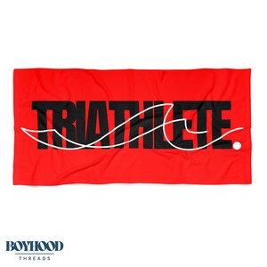 May include: A red beach towel with the word "TRIATHLETE" in large black letters. A white line design is overlaid on the text. The towel is rectangular and has the "BOYHOOD THREADS" logo in the bottom left corner.