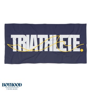 May include: Navy blue rectangular towel with the word "TRIATHLETE" in large white block letters. A yellow line weaves through the letters, and a small yellow dot is at the end. The brand "BOYHOOD THREADS" is at the bottom.