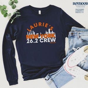 May include: Navy blue long-sleeve shirt with a white and orange graphic of a New York City skyline. The text "Laurie's New York 26.2 Crew" is printed on the shirt.
