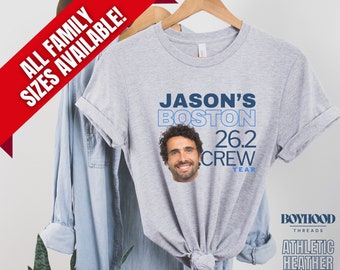 Könnte beinhalten: Graues T-Shirt mit einer blauen Grafik, auf der "Jason's Boston 26.2 Crew Year" steht, und einem Foto eines Mannes in der Mitte. Das Shirt hängt an einem Kleiderbügel und hat unten den Text "Boyhood Threads Athletic Heather".