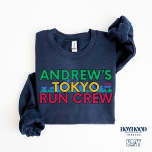 May include: Navy blue sweatshirt with the text "Andrew's Tokyo 2025 Run Crew" in colorful block letters. The text is centered on the sweatshirt.
