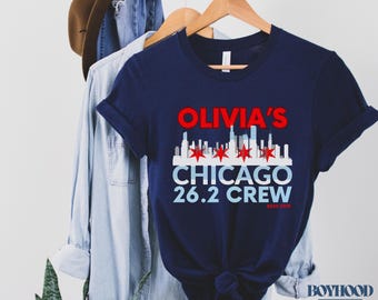 May include: Navy blue t-shirt with the text "Olivia's Chicago 26.2 Crew" in red and white. The design includes a skyline graphic and red stars. The shirt is displayed on a hanger with other clothing.