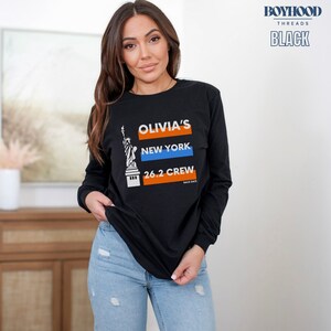 May include: Black long-sleeve t-shirt with a graphic of the Statue of Liberty and the text "Olivia's New York 26.2 Crew Race Date".