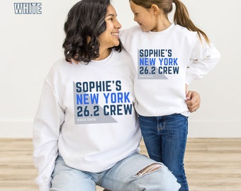 May include: White crew neck sweatshirts with the text "Sophie's New York 26.2 Crew" in blue and gray. The text "Race Date" is below the main text.