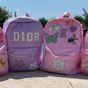 May include: Four pink and purple backpacks with various patches and embellishments. The backpacks have the names "ISLEE", "DIOR", "E", and "H" on them.