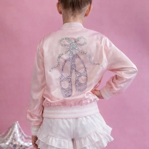 Puede incluir: Chaqueta bomber de satén rosa claro con un diseño de zapatillas de ballet de pedrería en la espalda. La chaqueta tiene ribetes a rayas blancas y rosas. El conjunto incluye shorts blancos con volantes.
