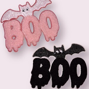 Może przedstawiać: Dwie dekoracje na Halloween. Jedna jest różowa z białym nietoperzem i napisem "BOO". Druga jest czarna z czarnym nietoperzem i napisem "BOO". Obie mają błyszczące wykończenie.
