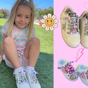 Könnte beinhalten: Ein junges Mädchen trägt ein pinkfarbenes und blaues Batik-Shirt und weiße Sneaker mit bunten Schnürsenkeln. Die Sneaker haben ein Sternendesign und den Text "CCDB" an der Seite.