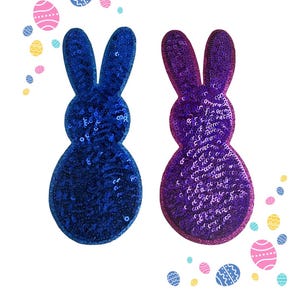 Könnte beinhalten: Zwei paillettenbesetzte Osterhasen-Dekorationen in Blau und Lila, mit dekorativen Ostereiern in Pastellfarben verstreut. Die Hasen sind in vertikaler Pose, mit langen Ohren und rundem Körper.