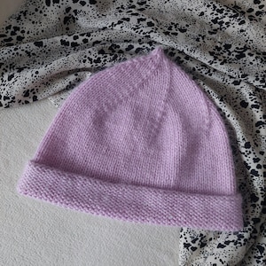 Puede incluir: Un gorro de punto lila claro con un borde doblado. El gorro está hecho de un hilo suave y texturizado. El fondo incluye una superficie blanca y una tela estampada con manchas negras y blancas.