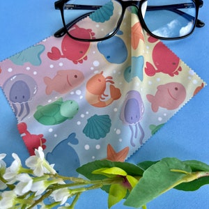 May include: A microfiber cleaning cloth with a sea life pattern in shades of pink, blue, green, and yellow. The cloth is folded and a pair of black framed glasses are lying on top of it. The glasses have a blue tint and reflect the sky.