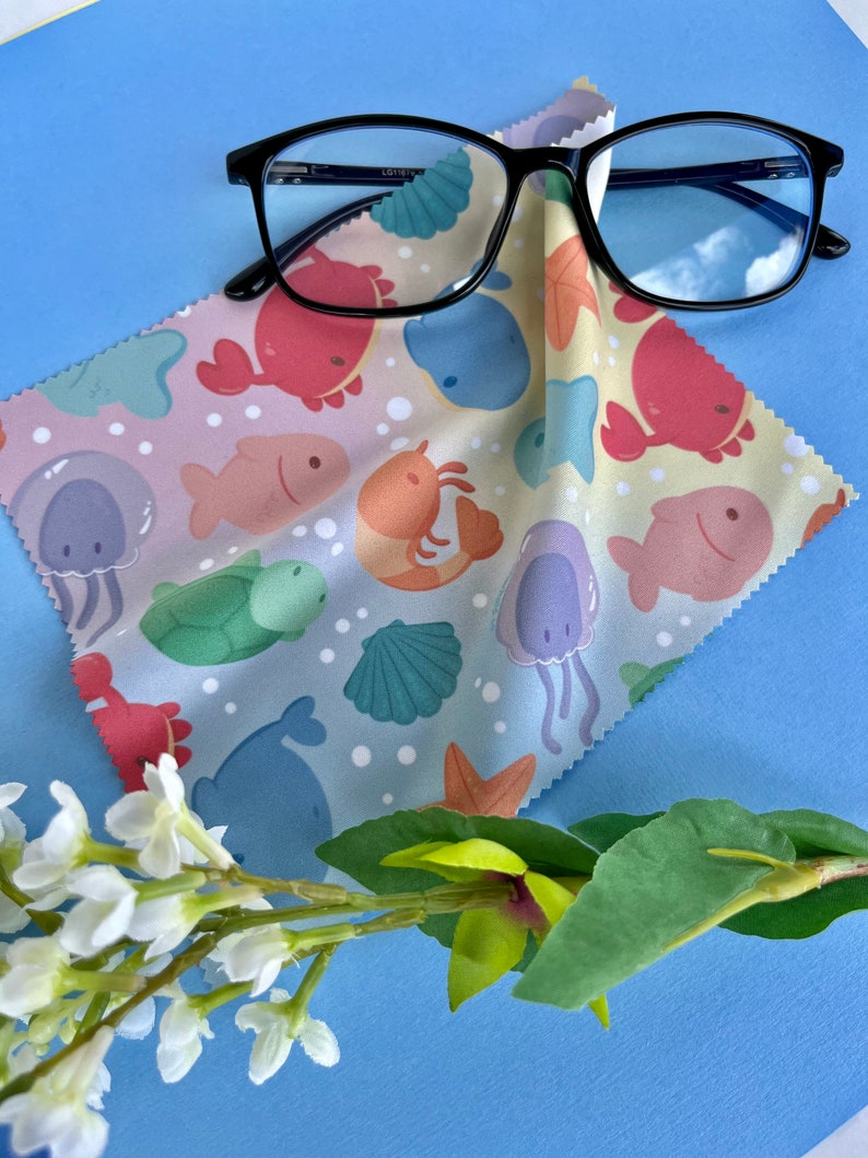May include: A microfiber cleaning cloth with a sea life pattern in shades of pink, blue, green, and yellow. The cloth is folded and a pair of black framed glasses are lying on top of it. The glasses have a blue tint and reflect the sky.