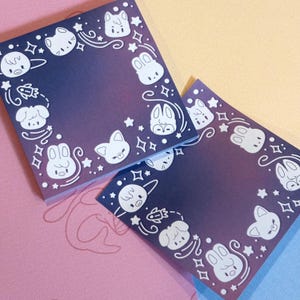 May include: Two square sticky note pads featuring a dark purple to light purple gradient background. The border of each note is decorated with white line art illustrations of various small animal faces, stars, and swirling patterns.