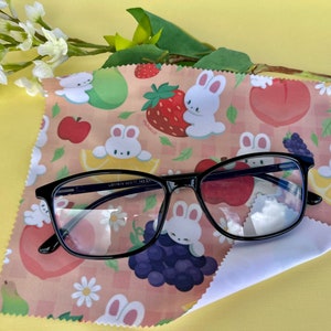 May include: A black framed pair of glasses resting on a pink and white microfiber cleaning cloth with a pattern of white bunnies, strawberries, peaches, lemons, and grapes. The cloth has a checkered pattern.