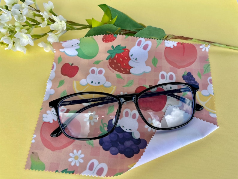 May include: A black framed pair of glasses resting on a pink and white microfiber cleaning cloth with a pattern of white bunnies, strawberries, peaches, lemons, and grapes. The cloth has a checkered pattern.