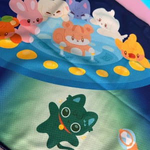 May include: A close-up of a blue and green fabric with cartoon animal illustrations. The design features a variety of cute characters, including a pink bunny, a white seal, and a green cat. The background has a starry night sky theme.