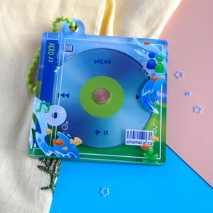 K&ouml;nnte beinhalten: Ein klarer Acryl-Schl&uuml;sselanh&auml;nger mit einem CD-Design in Gr&uuml;n und Blau. Der Schl&uuml;sselanh&auml;nger hat eine gr&uuml;ne Perlenkette und enth&auml;lt den Text "AERO" und "MENU". Kleine orangefarbene Fische und Blasen verzieren das Design. Der Text "ohwhale.co" ist ebenfalls sichtbar.
