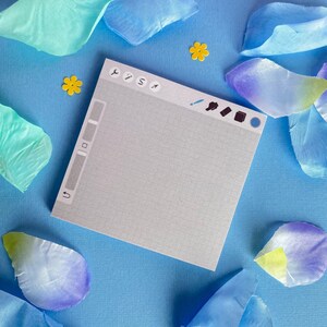 May include: A square notepad featuring a light gray grid pattern and a digital interface design at the top with various tool icons. The notepad is surrounded by scattered blue and teal fabric flower petals and small yellow flower-shaped confetti.