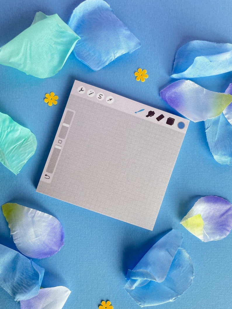 May include: A square notepad featuring a light gray grid pattern and a digital interface design at the top with various tool icons. The notepad is surrounded by scattered blue and teal fabric flower petals and small yellow flower-shaped confetti.