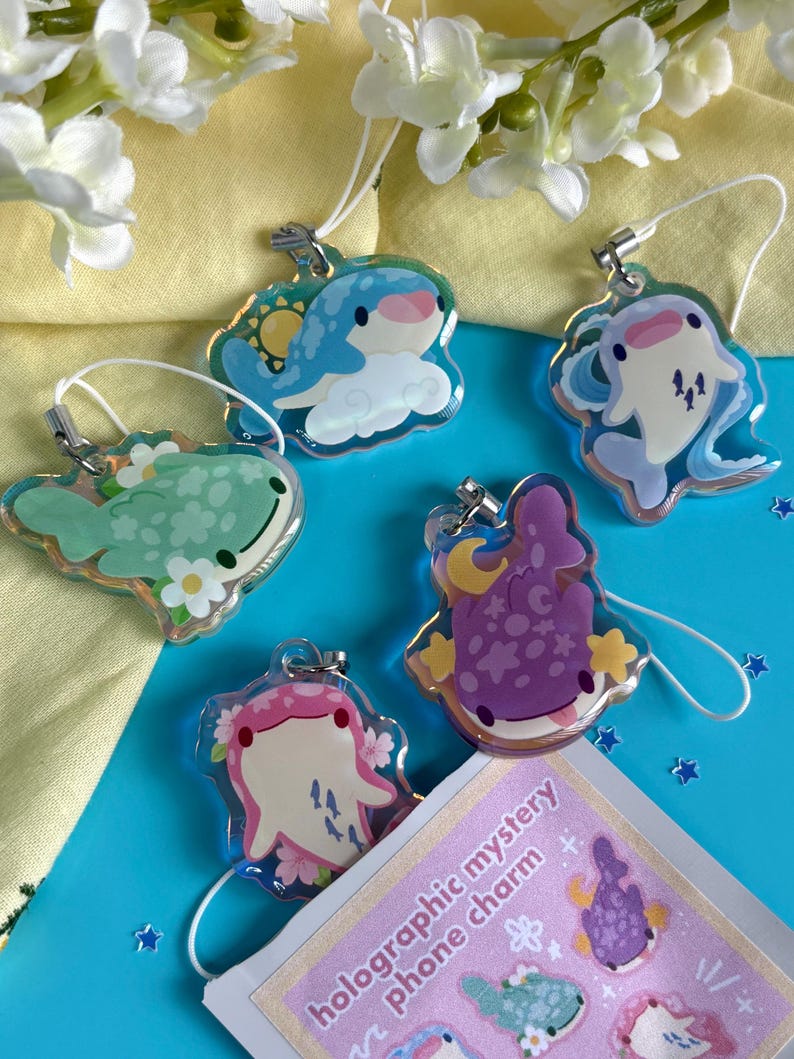 May include: A collection of holographic mystery phone charms featuring various sea creatures. The charms are in the shape of whales, dolphins, and other marine life, with colorful designs and attached lanyards. The charms are on a blue surface.