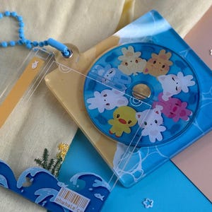 May include: A clear acrylic keychain with a blue and yellow theme. The keychain features a circular design with cartoon animals and a wave-shaped accent. The text "ohwhale.co" is visible on the wave accent. A blue beaded chain is attached.