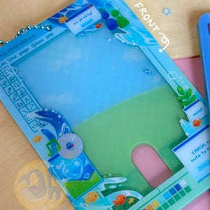 May include: A clear, blue card holder with a whimsical design. The holder features a landscape scene with fish, water, and text that reads "so pretty!" and "wow!". The front of the holder is labeled "FRONT".