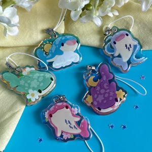 May include: Five clear acrylic charms with cartoon whale designs. The charms feature various colors, including blue, green, pink, and purple, with floral and celestial accents. Each charm has a silver-colored clasp and a white string.
