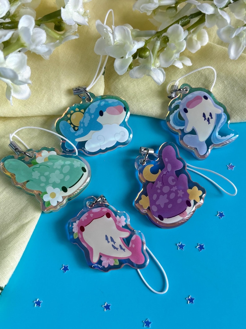 May include: Five clear acrylic charms with cartoon whale designs. The charms feature various colors, including blue, green, pink, and purple, with floral and celestial accents. Each charm has a silver-colored clasp and a white string.