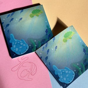 May include: Two blue and green patterned sticky notes with a sea turtle and a whale shark illustration. The notes are on a pink and blue background.