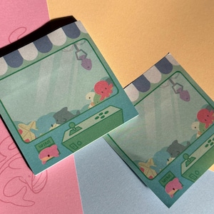 May include: Two square sticky notes featuring a claw machine illustration. The design includes a blue and white striped awning, a green control panel with a joystick, and various sea creature plushies inside. The word WIN is printed on the front.