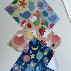 May include: Three different patterned microfiber cloths. The first cloth has a light blue background with a pattern of sea creatures, including a whale, jellyfish, and starfish. The second cloth has a peach background with a pattern of bunnies and fruit, including strawberries, apples, and lemons. The third cloth has a dark blue background with a pattern of planets, stars, and a cat astronaut.
