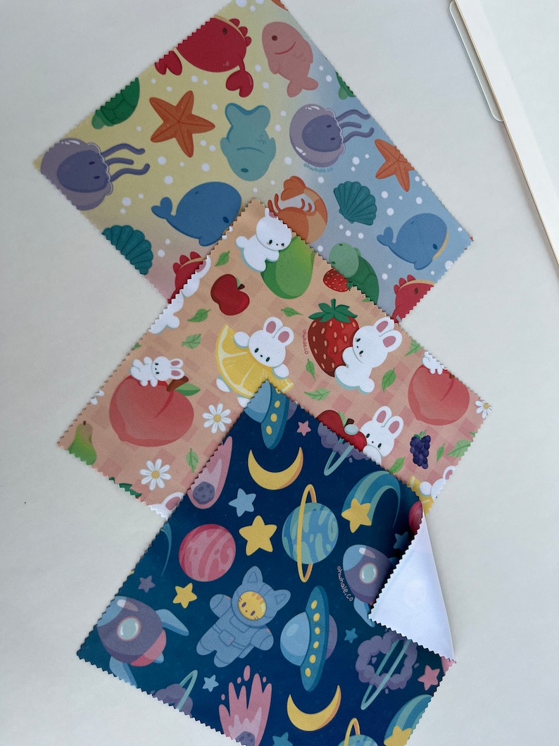 May include: Three different patterned microfiber cloths. The first cloth has a light blue background with a pattern of sea creatures, including a whale, jellyfish, and starfish. The second cloth has a peach background with a pattern of bunnies and fruit, including strawberries, apples, and lemons. The third cloth has a dark blue background with a pattern of planets, stars, and a cat astronaut.