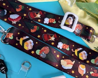 Puede incluir: Un cordón rojo y negro con un patrón de sushi y estrellas. El cordón tiene una hebilla de plástico blanco y un portagafete de plástico transparente. El cordón está sobre una superficie azul.