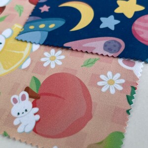 May include: Two fabric swatches with different patterns. The top swatch is navy blue with white stars, a yellow crescent moon, and a pink and purple planet. The bottom swatch is peach with white daisies, green leaves, and a pink peach with a white bunny.
